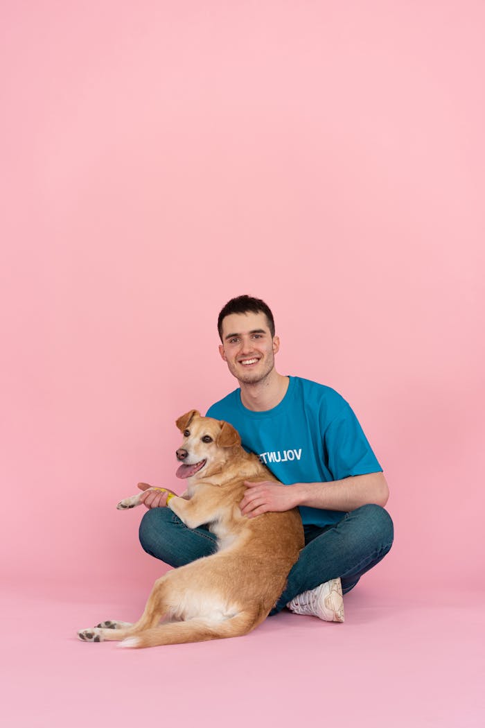 A young man smiles while sitting with a dog against a pink background, depicting joy and companionship.